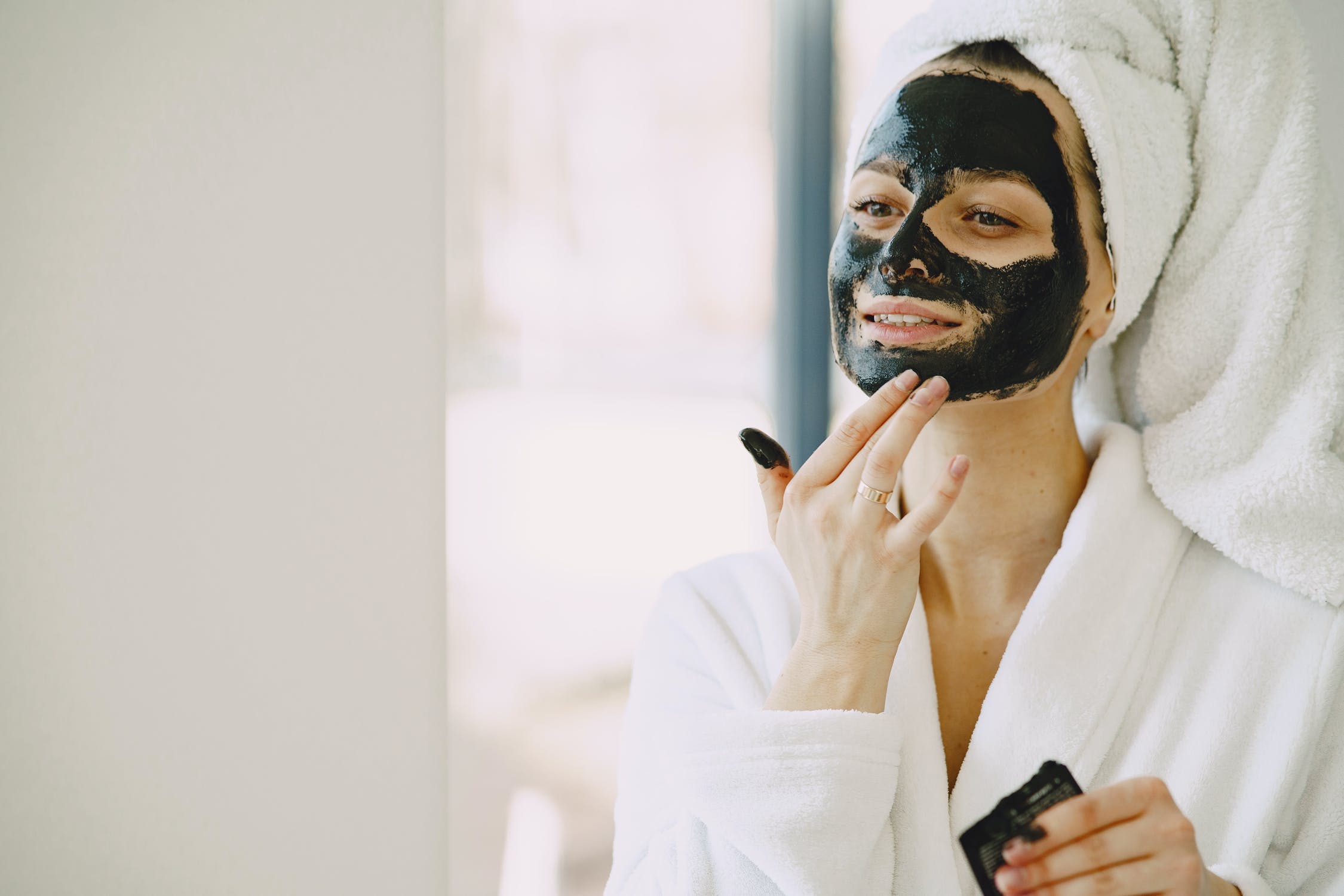 mulher vestindo roupão e toalha na cabeça aplicando máscara facial de argila preta