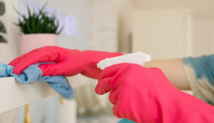 foto mostra mãos usando luvas, limpando uma superfície com frasco spray e pano de limpeza