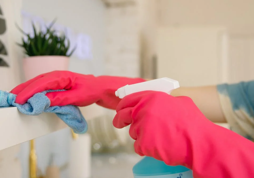 foto mostra mãos usando luvas, limpando uma superfície com frasco spray e pano de limpeza