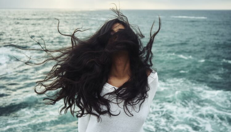 A imagem mostra uma mulher com cabelos longos ao vento em frente ao mar