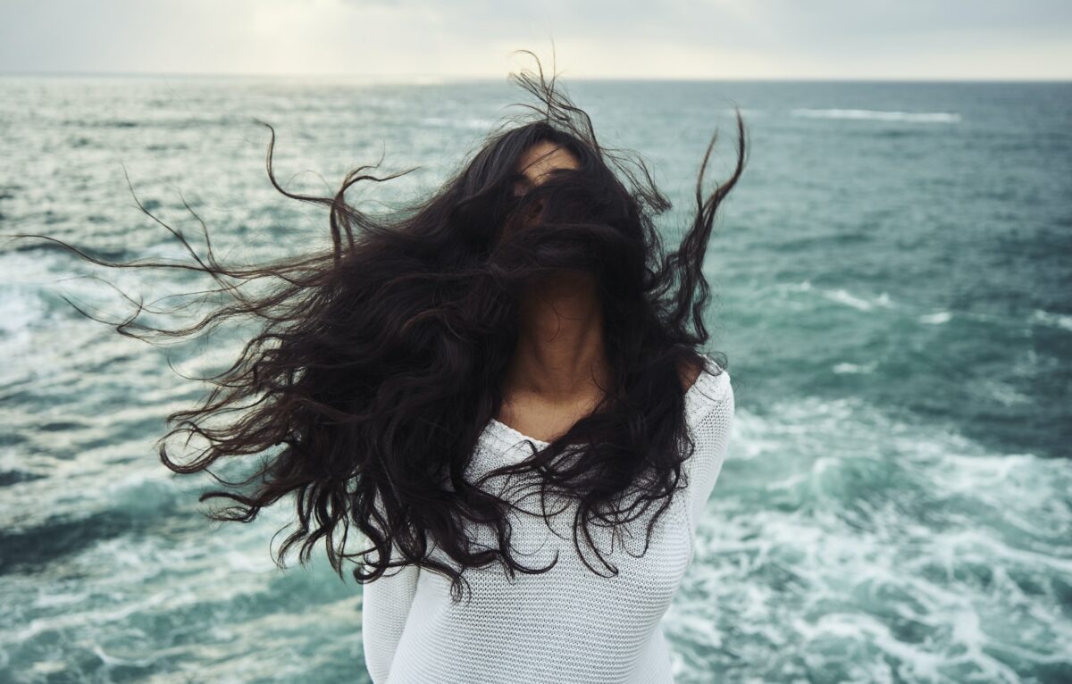 A imagem mostra uma mulher com cabelos longos ao vento em frente ao mar