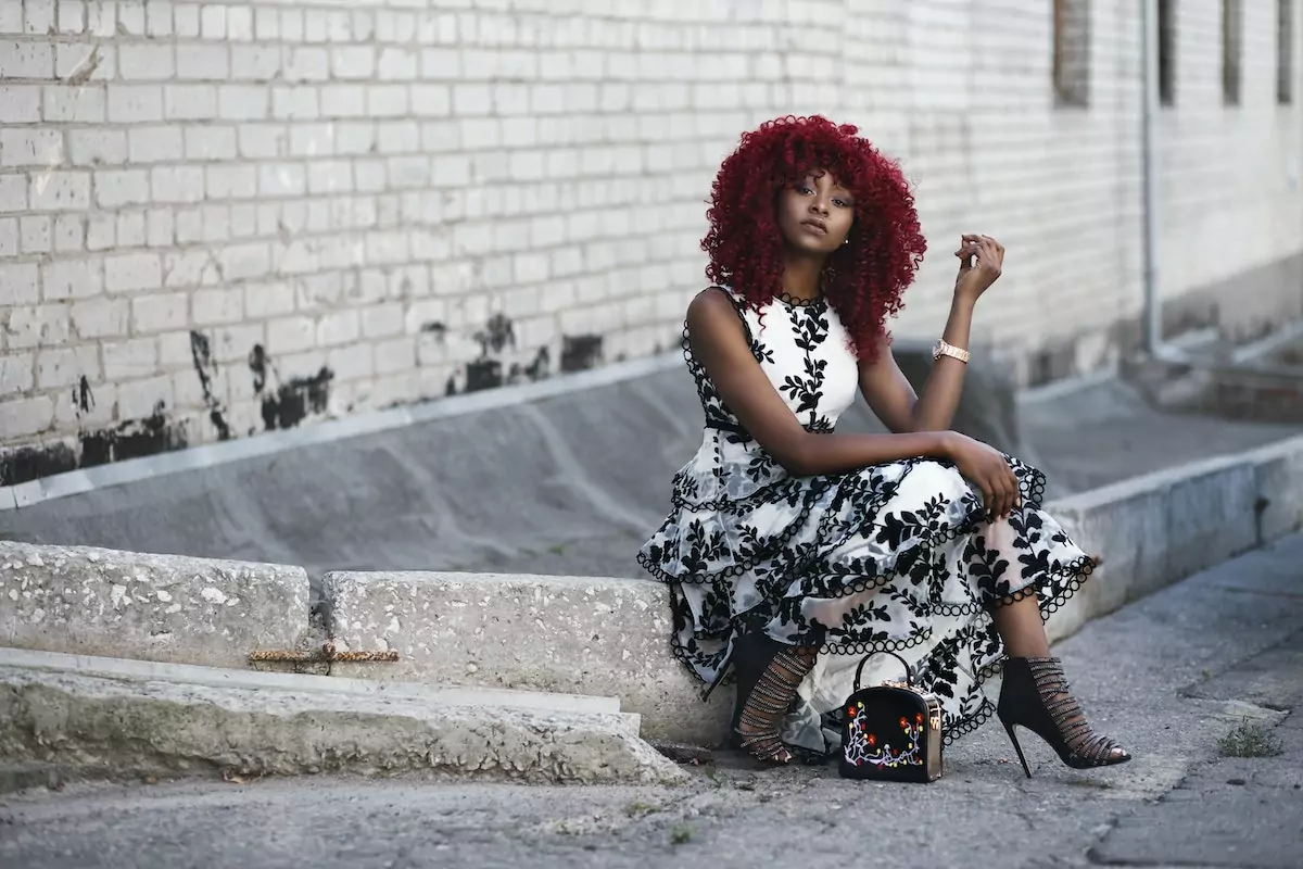 mulher negra de cabelo cacheado vermelho com vestido branco e preto e salto alto. Ela está sentada em uma calçada posando para foto