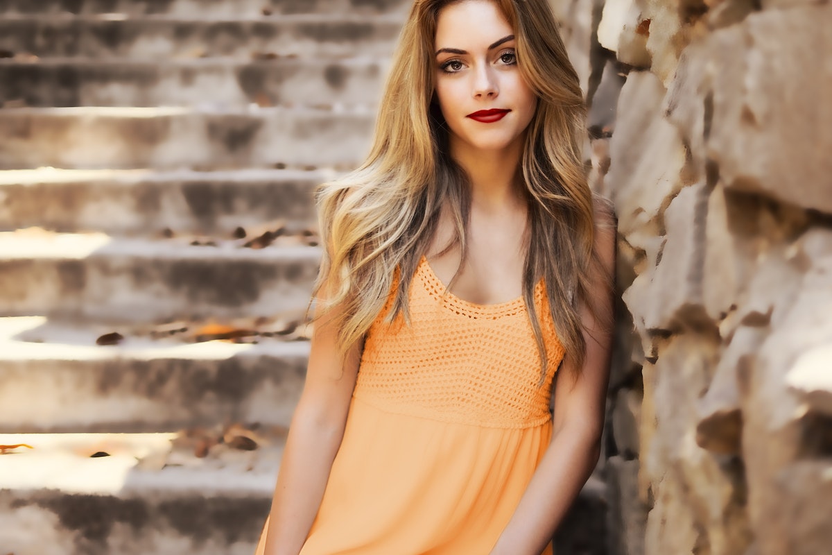 Mulher na rua com cabelo claro e vestido laranja encostada em uma parede e, atrás dela, há uma escada