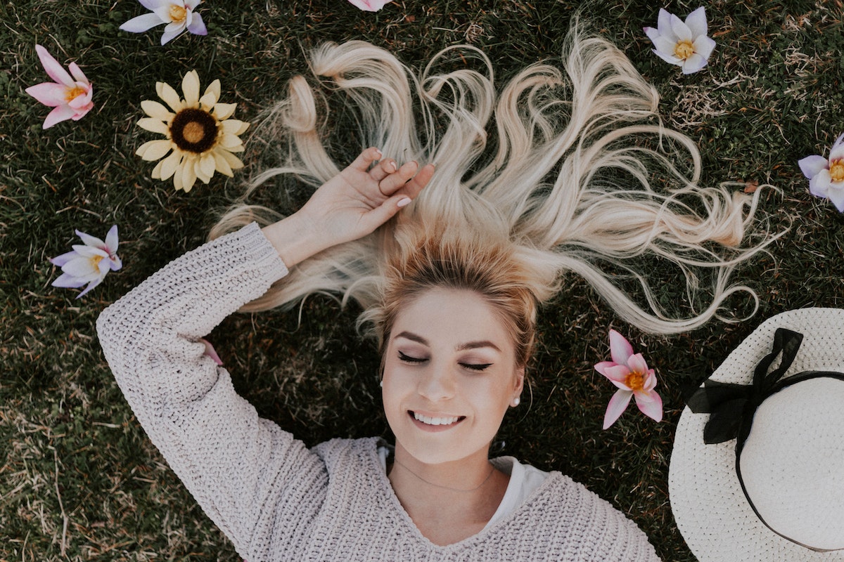 mulher de cabelo loiro sorrindo, deitada na grama e, ao seu redor, há um chapéu branco e diversas flores amarelas, azuis e rosas