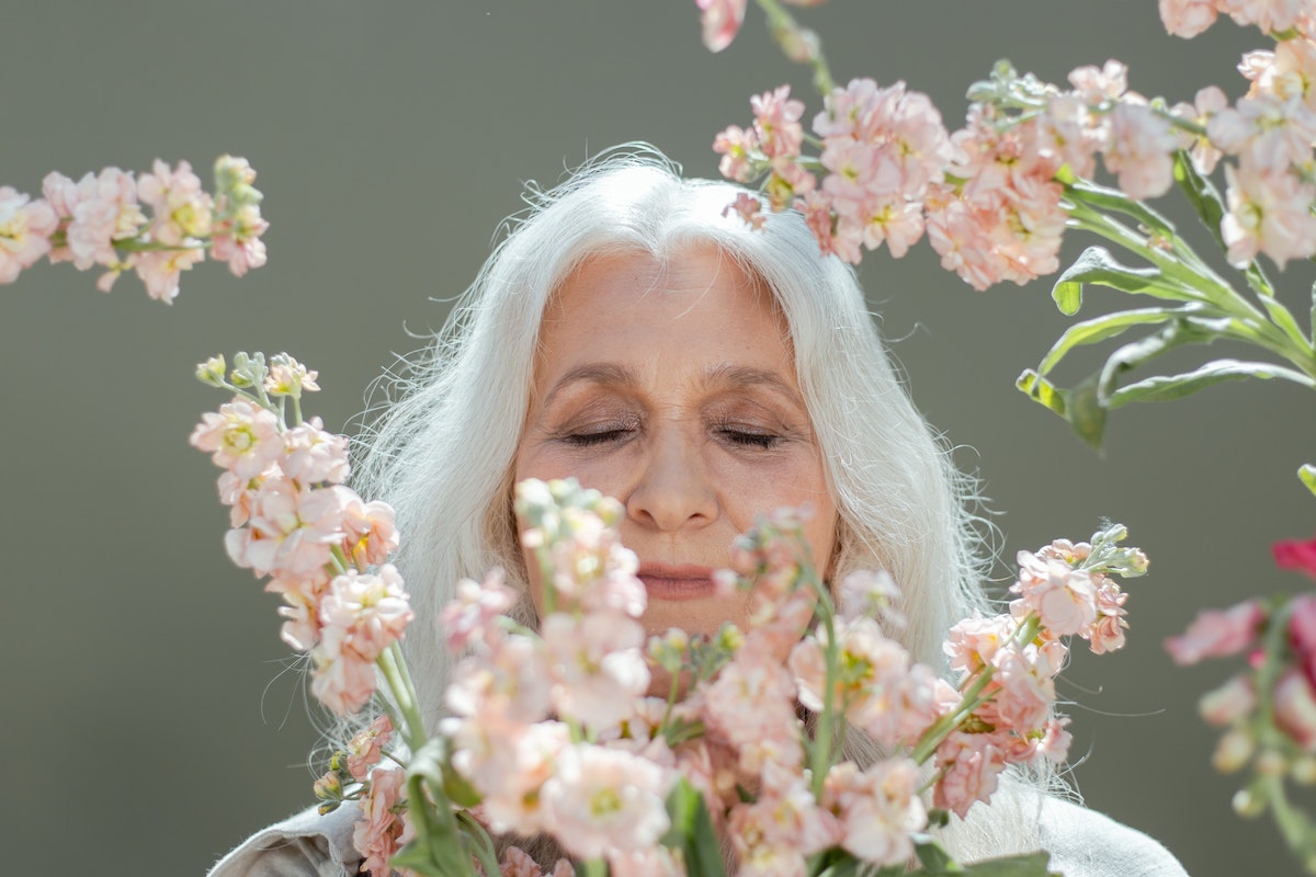 mulher com cabelo grisalho solto posando para foto. Seu rosto está próximo a ramos de plantas com flores rosas