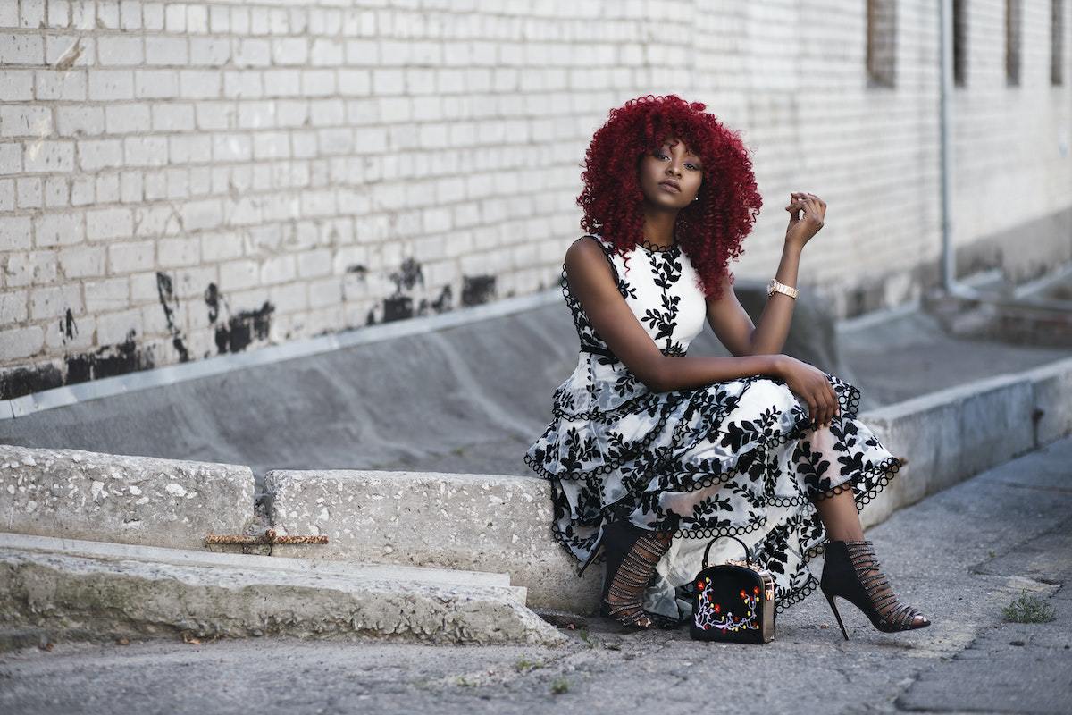 mulher negra de cabelos ruivos cacheados posando para foto, sentada em um paralelepípedo de calçada