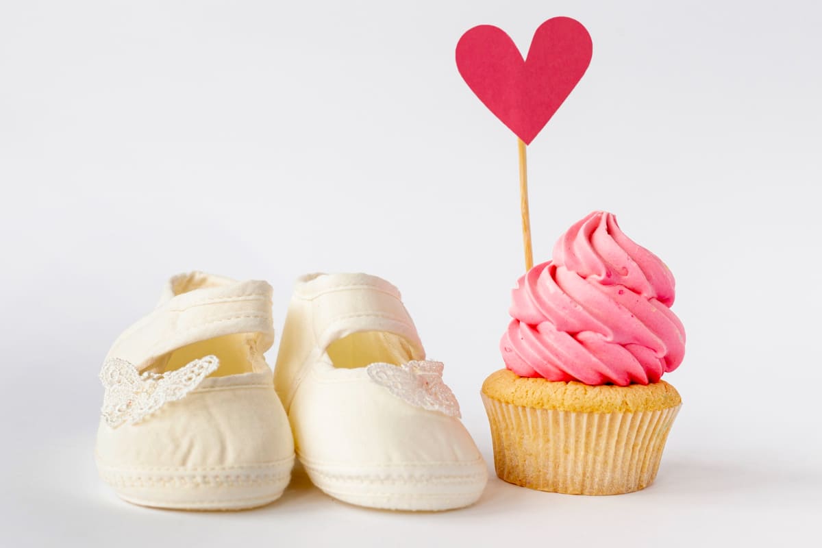 imagem de um sapatinho de bebê branco e, do lado, um cupcake rosa com uma imagem de coração como item decorativo