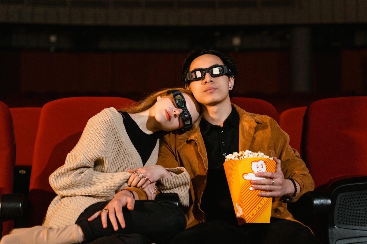 homem e mulher sentados em sala de cinema com óculos 3D. O homem está segurando um balde de pipoca, enquanto a mulher encosta sua cabeça em seu ombro e segura o braço do companheiro
