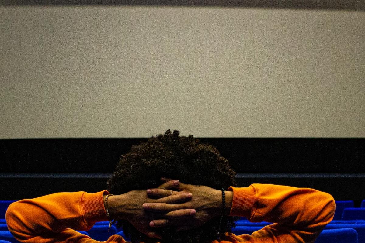homem negro de cabelo cacheado usando moletom laranja, sentado em frente a uma tela de cinema