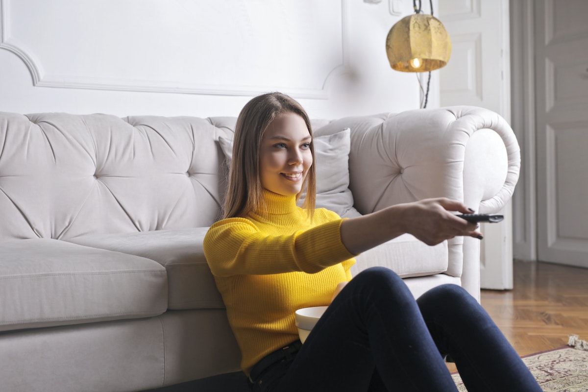 Mulher loira sentada no chão encostada no sofá com controle de TV na mão
