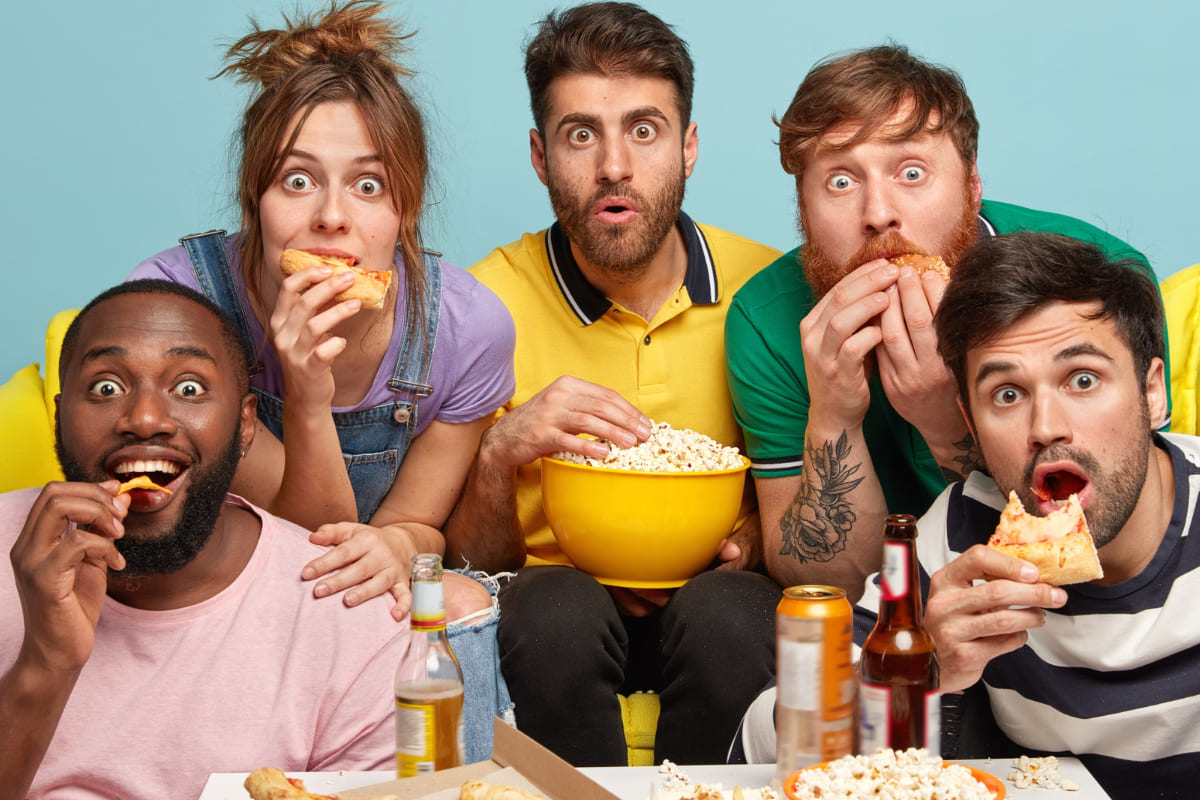 Grupo de 5 pessoas sentadas em um sofá comendo pipoca, chips e pizza. Todos estão com expressão de surpresa
