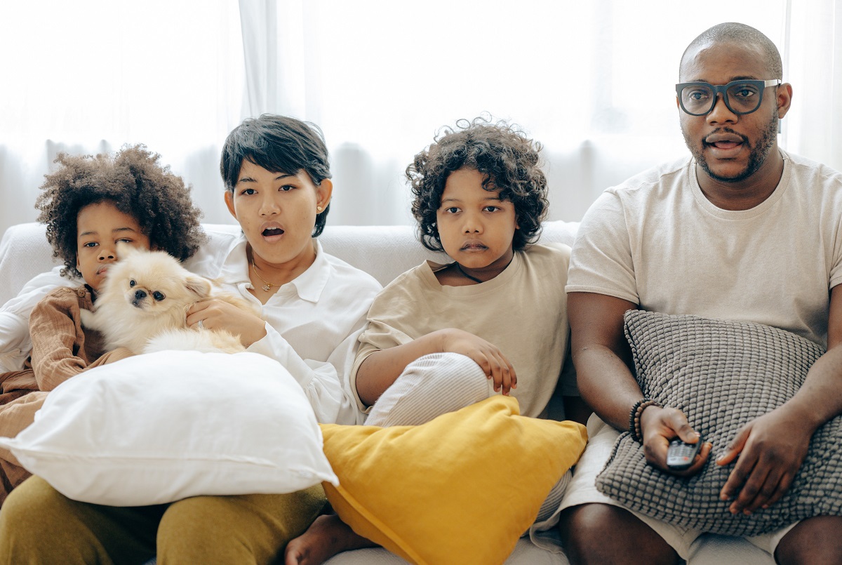 dois adultos e duas crianças de pele negra sentados em um sofá branco com almofadas de diversas cores