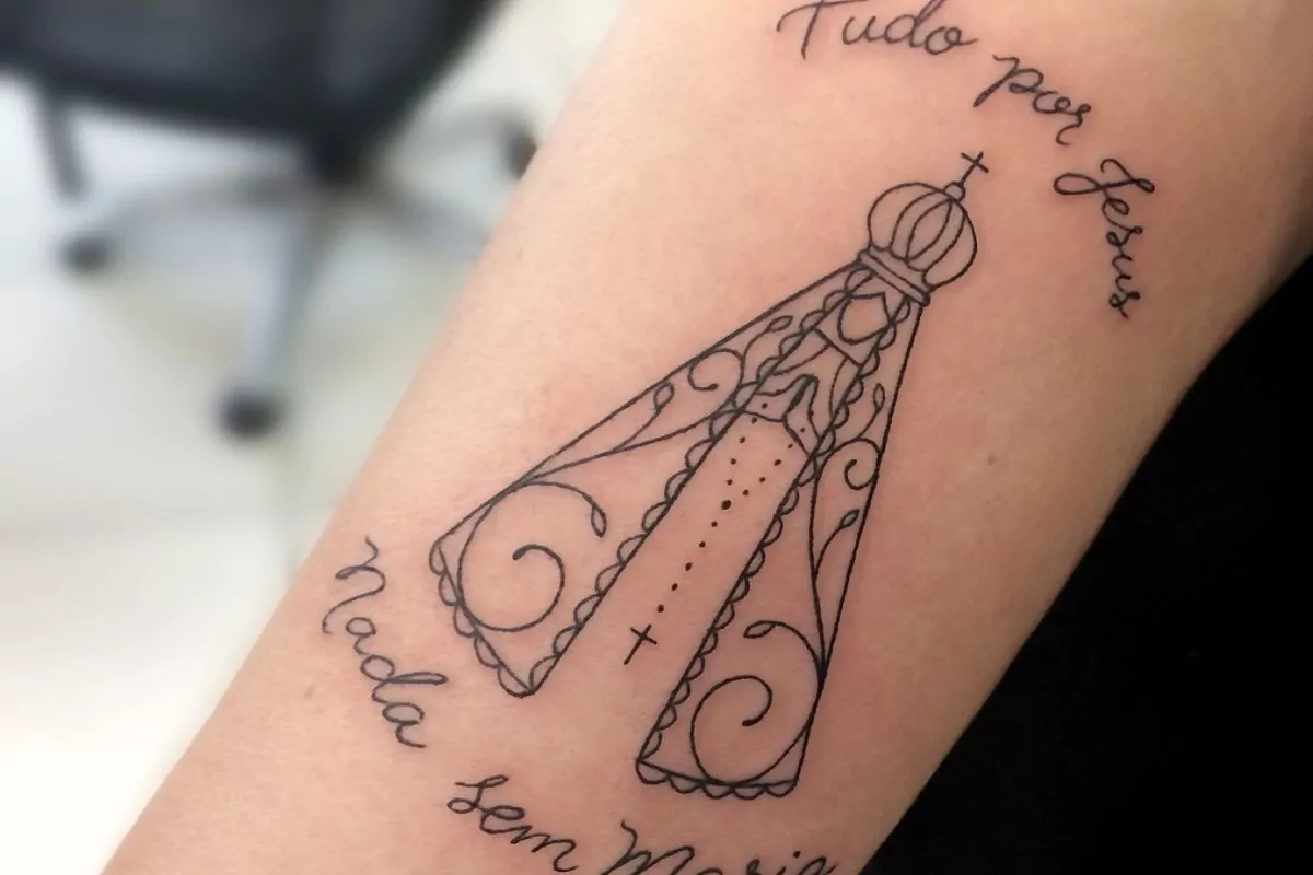Imagem de um braço com a tatuagem de Nossa Senhora de Aparecida com a frase "tudo por Jesus e nada sem Maria"