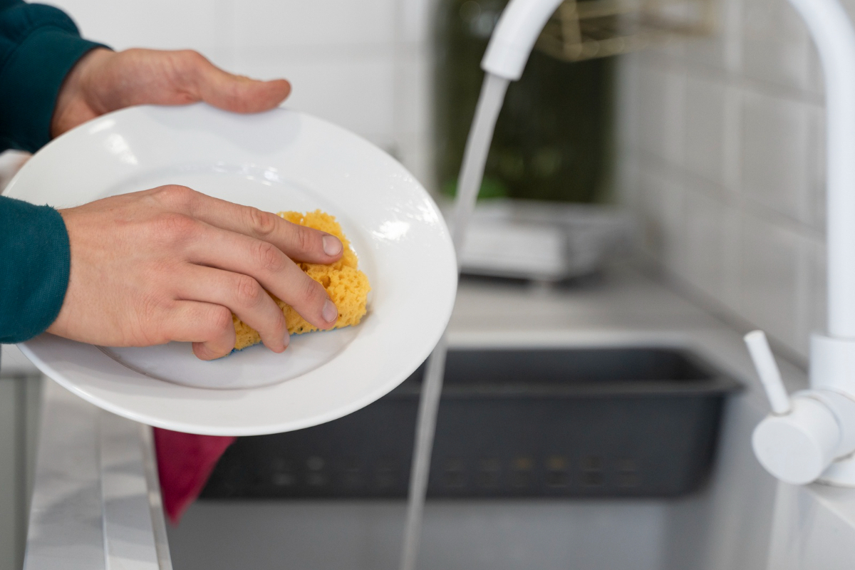 Imagem de uma mão feminina lavando um prato com esponja na pia sob a água