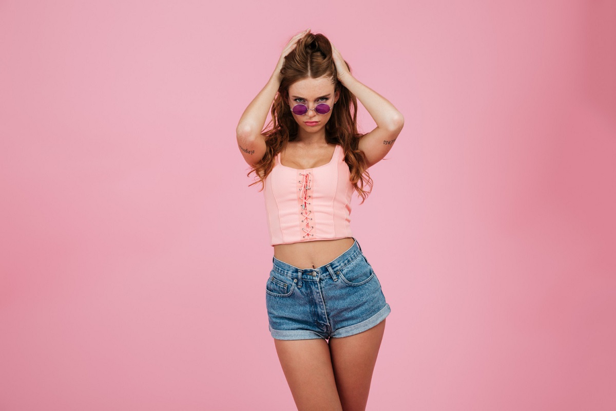 Mulher diante de parede rosa com look composto por shorts, blusa rosa e óculos de sol