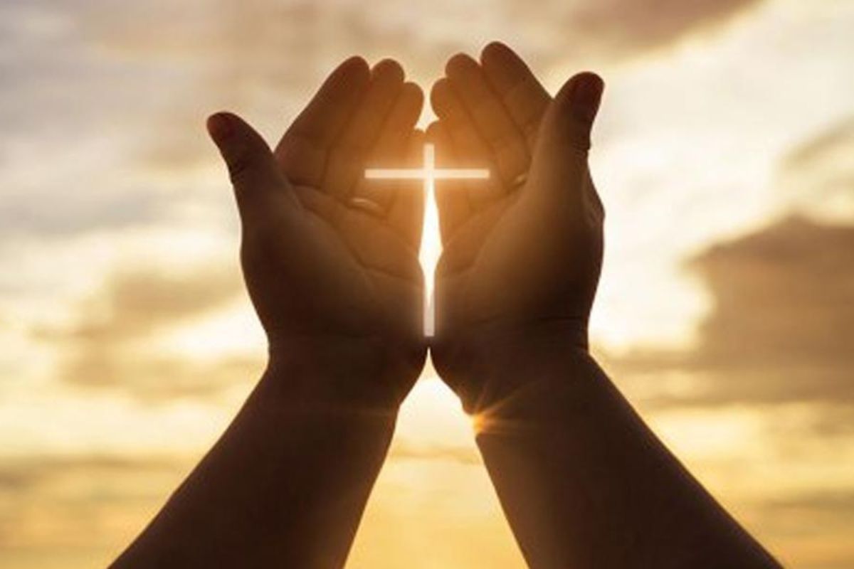Pessoa com mãos erguidas e juntas diante de por do sol com montagem que forma uma cruz entre as suas palmas