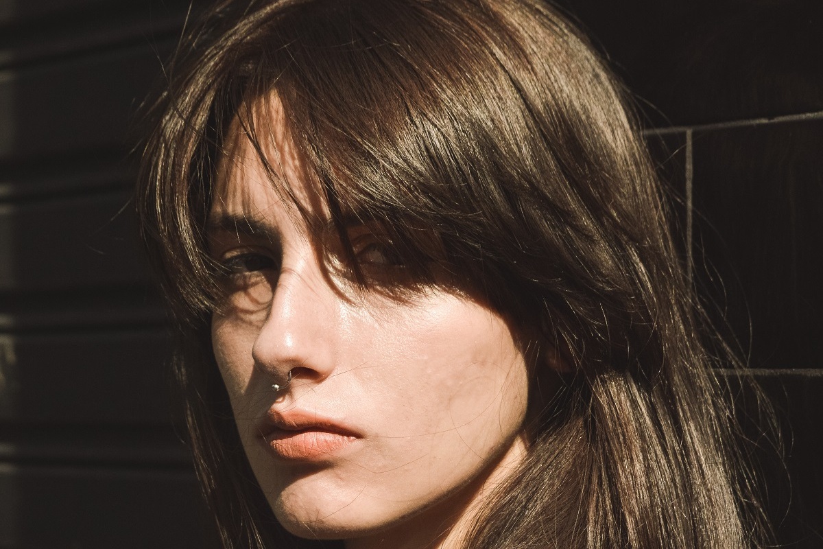 Foto com foco em rosto de mulher com piercing no nariz e cabelo liso com franja estilo retrô anos 80 e parede de tijolos em desfoco ao fundo da imagem