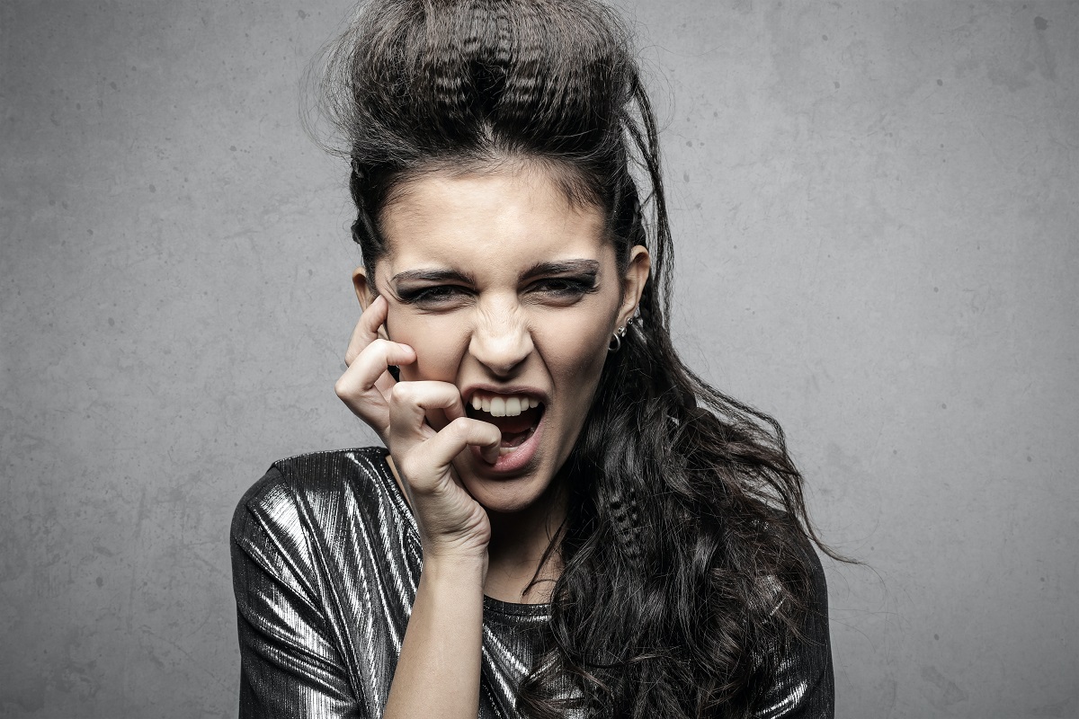 Imagem de mulher com aparência de roqueira com mão no rosto enquanto morde as unhas e maquiagem bem pesada com ar gótico e penteado que remete aos anos 80