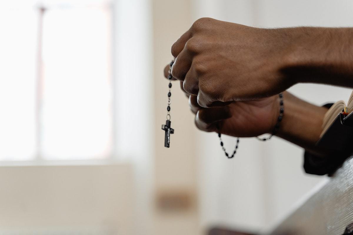 Pessoa aparentemente de joelhos com mãos estendidas à frente do corpo segurando terço em local análogo à igreja fazendo suas orações