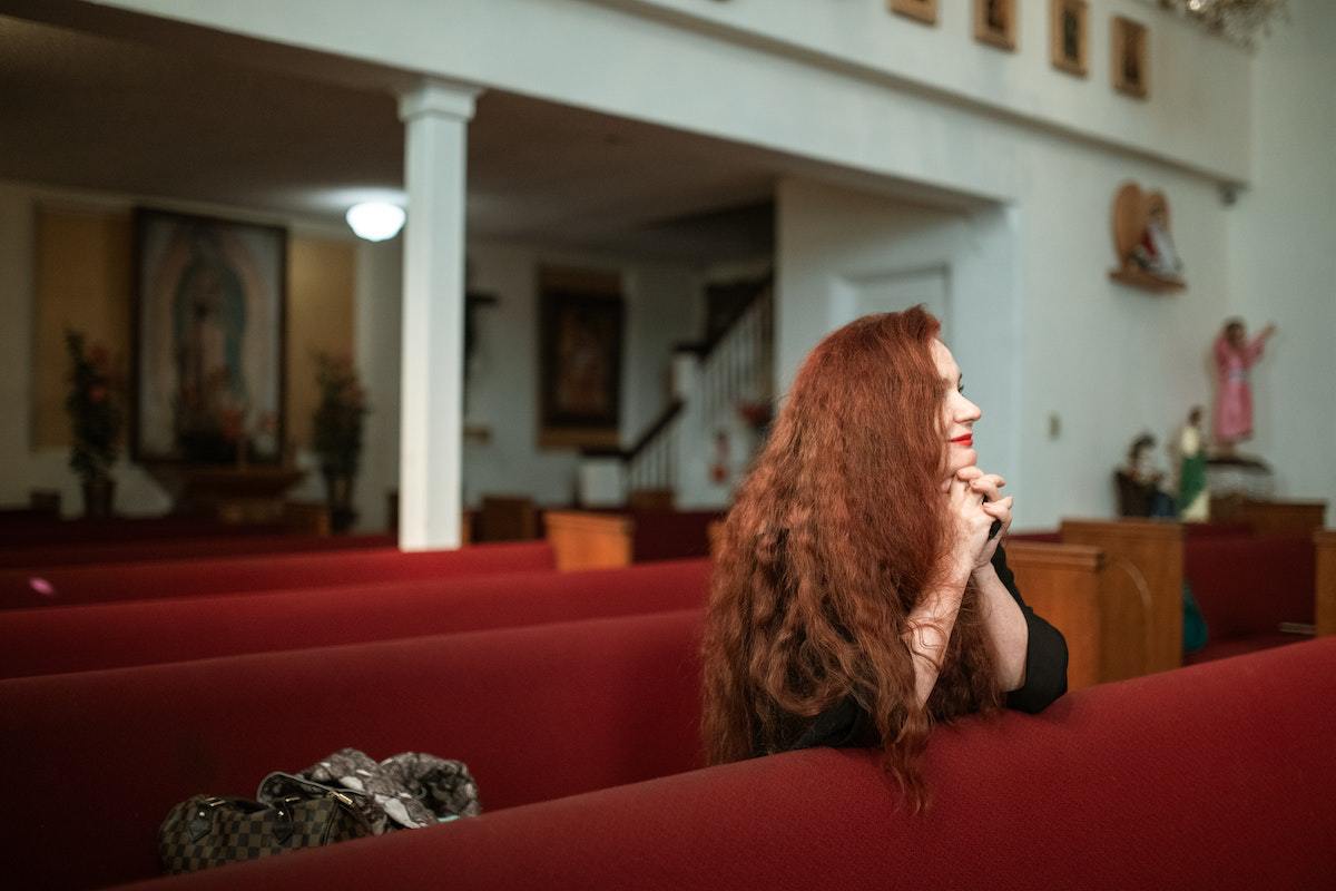 Mulher em igreja pequena com bancos vermelhos ajoelhada em meio à fileiras destes fazendo sua oração olhando para frente para o altar e com mãos juntas