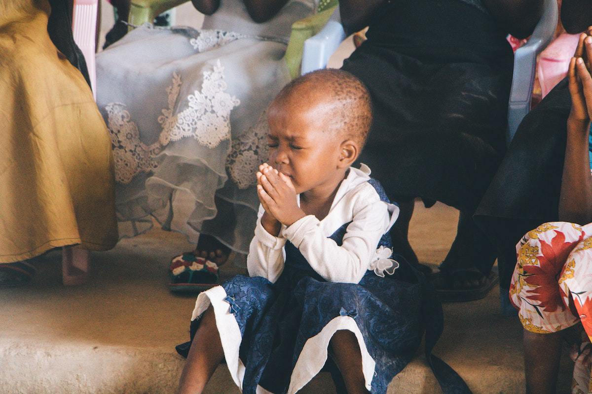 Pequena menina de cerca de cinco a seis anos de idade sentada no chão de igreja usando vestido azul escuro diante de senhoras sentadas em bancos com mãos unidas em sinal de oração
