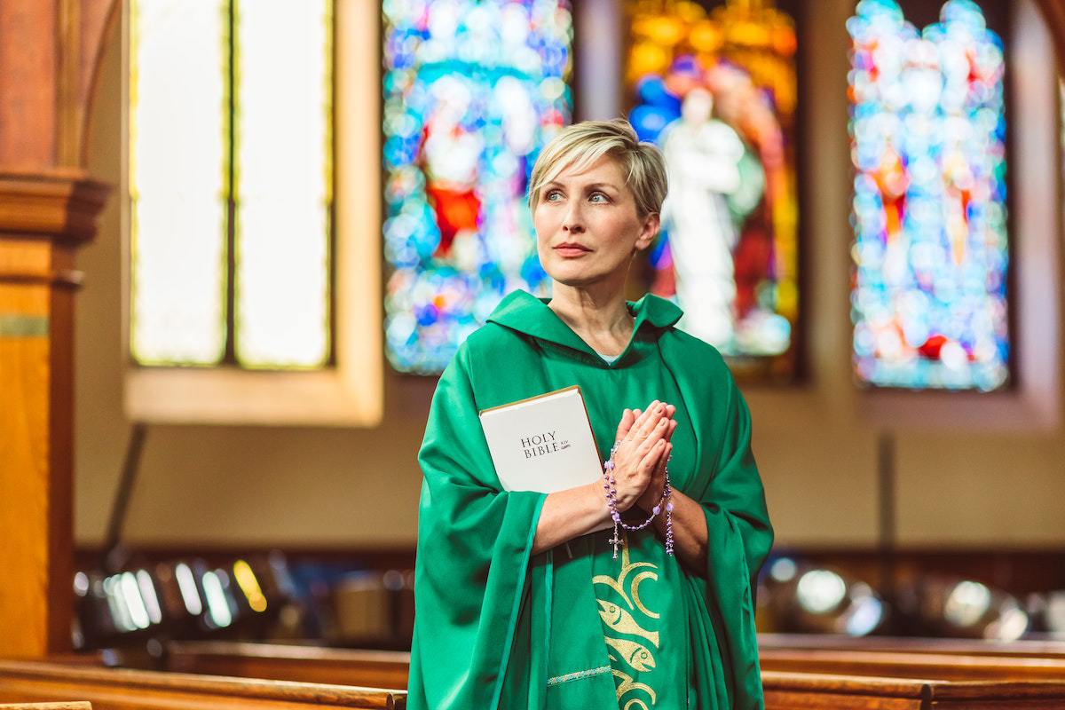 Mulher com túnica verde e mãos unidas em sinal de oração dentro de igreja e com bíblia de capa branca entre seus braços dentro de igreja