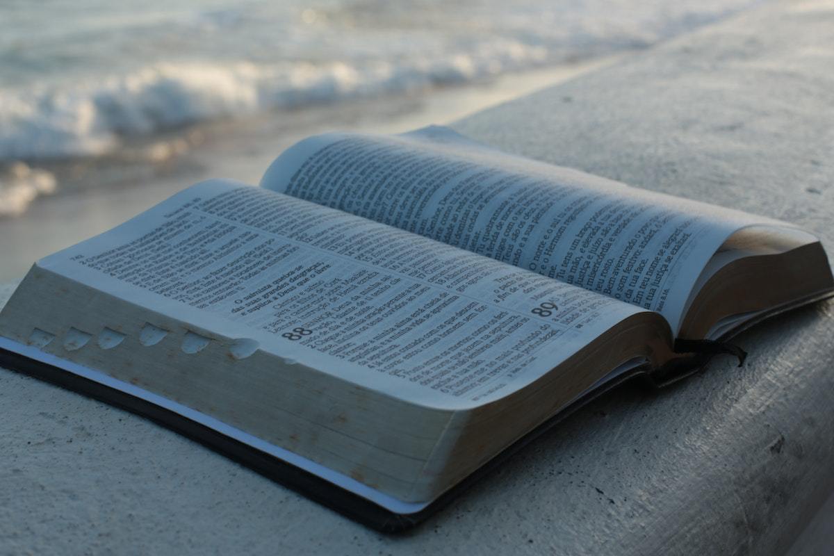 Bíblia aberta sobre pequena mureta de concreto na cor cinza que divide a orla da praia com imagem de pequena onda em desfoco se formando ao fundo da fotografia