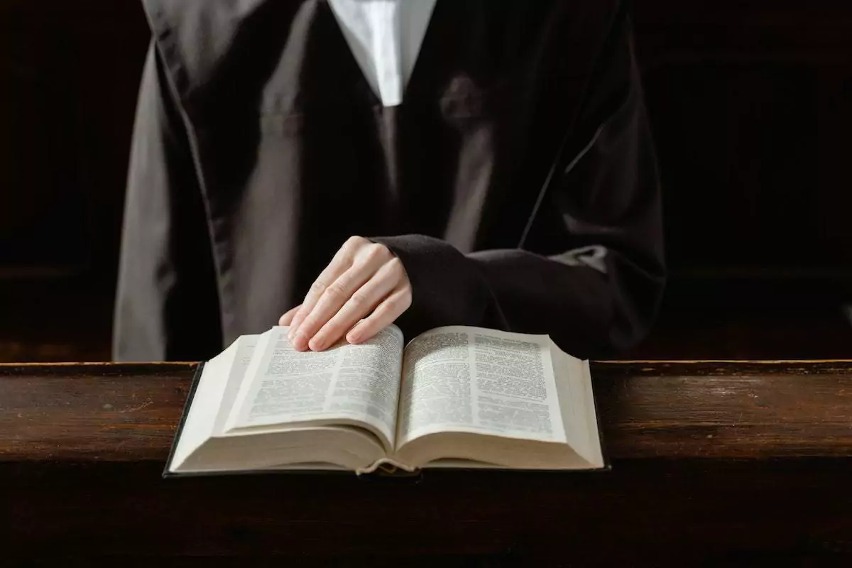 Pessoa usando grande túnica de mangas longas pretas com vestimenta branca por baixo folheando bíblia que está apoiada sobre superfície de madeira aparentemente em interior de igreja cristã