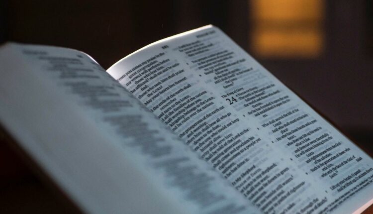 Bíblia cristã aberta sobre pequeno suporte de madeira fina com imagem de janela de vidro em desfoco ao fundo de foto que foi tirada em ambiente de pouca luminosidade