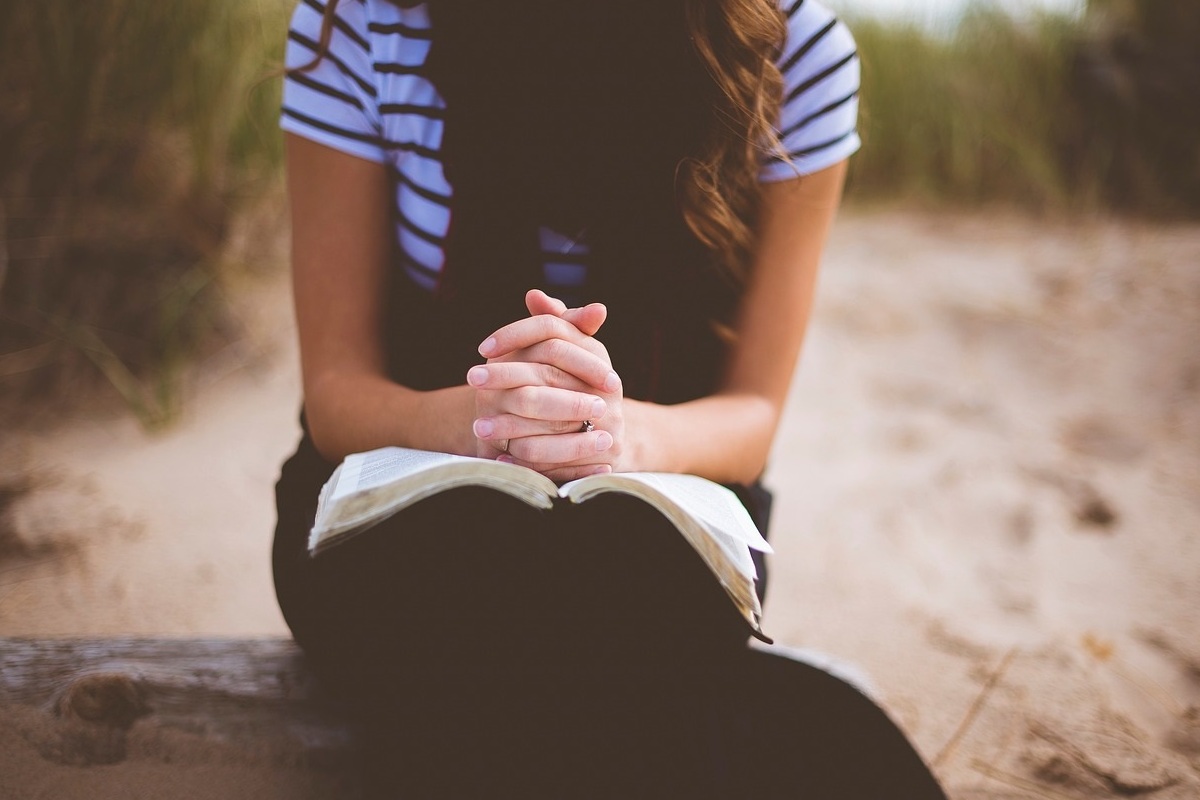 Mulher orando em ambiente natural com bíblia no colo e mãos com dedos entrelaçados