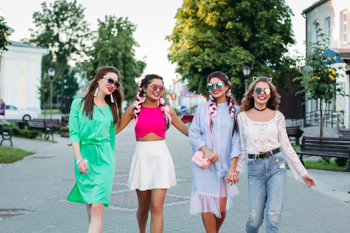 Quatro mulheres caminhando no meio de avenida estreita usando roupas leves e coloridas lado a lado sorridentes e com óculos de verão em moda dopamina