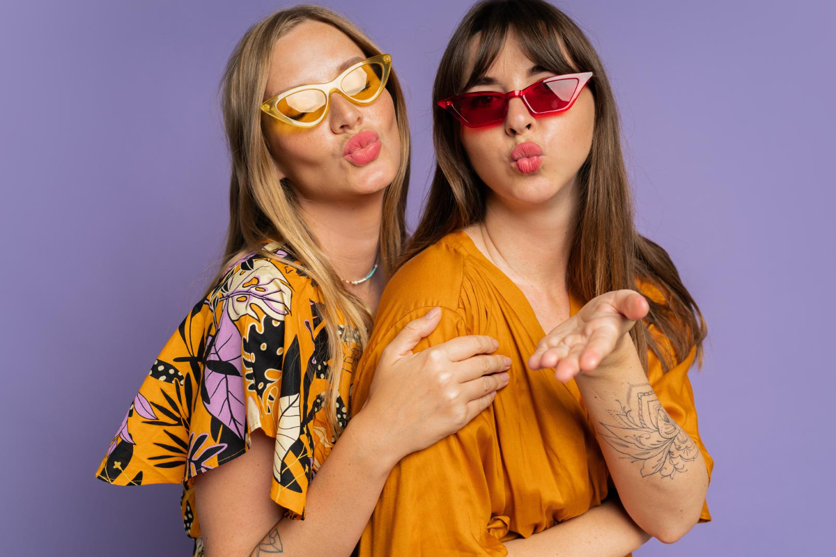 Duas mulheres diante de parede roxa fazem pose para foto com biquinhos de beijo usando óculos de verão meio pontiagudos, posicionadas meio de lado com uma por trás da outra colocando a mão em seu ombro
