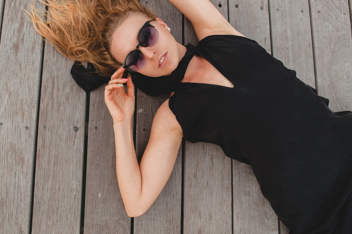 Mulher com vestido preto e óculos escuros de lente dégradé de preto e roxo deitada em piso de madeira com tábuas finas enquanto segura na perna direita do óculos