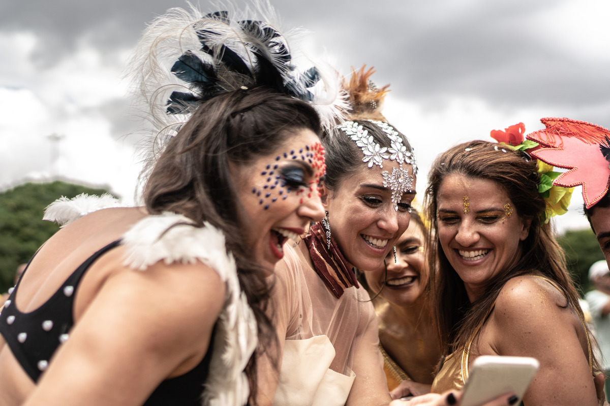 Três mulheres legendando post com frases de carnaval; estão todas fantasiadas, olhando para a tela de um telefone e sorrindo