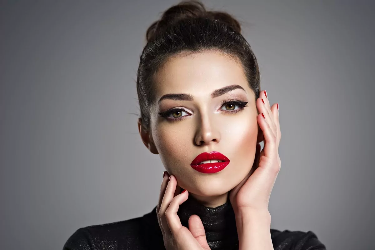 mulher morena com maquiagem forte e batom vermelho, posando com as mãos no rosto
